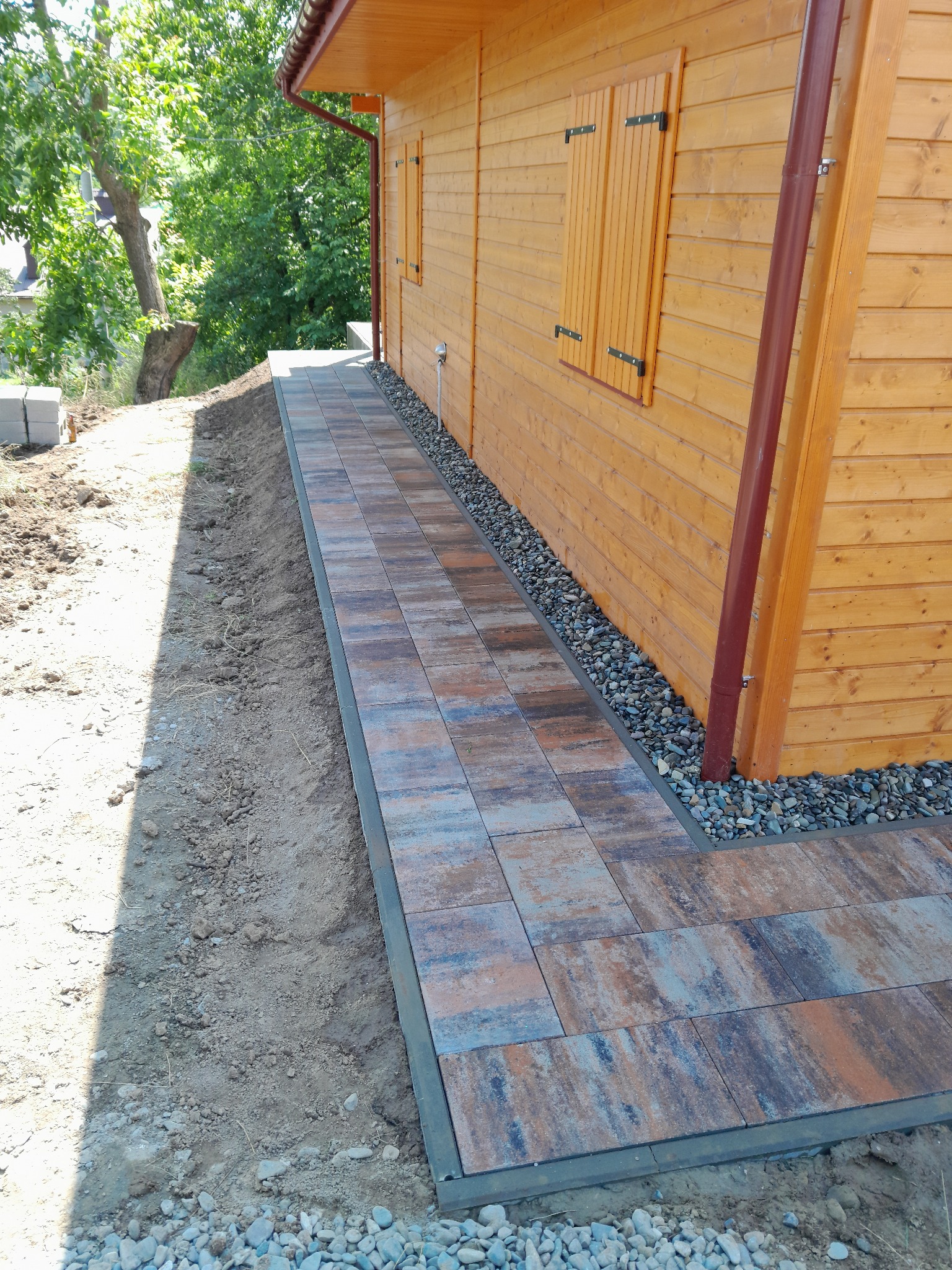 Ścieżka z kostki brukowej prowadząca wzdłuż drewnianego domku z brązowymi okiennicami, obok nasypu ziemi i kamieni ozdobnych. Ujęcie z perspektywy.