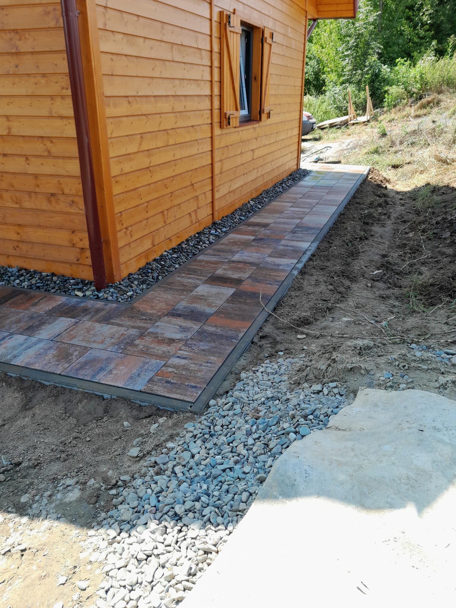 Ścieżka z kostki brukowej prowadząca do drewnianego domku, obrzeża wysypane drobnymi kamieniami. Ujęcie z boku budynku, widoczne okno z okiennicą.