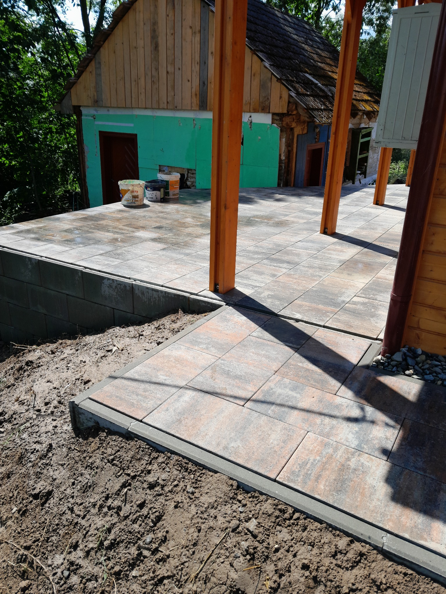 Ułożona kostka brukowa na tarasie z drewnianą konstrukcją, widoczne wiaderka z farbą i fragment budynku w tle. Kostka w odcieniach szarości i brązu, świeżo ułożona.