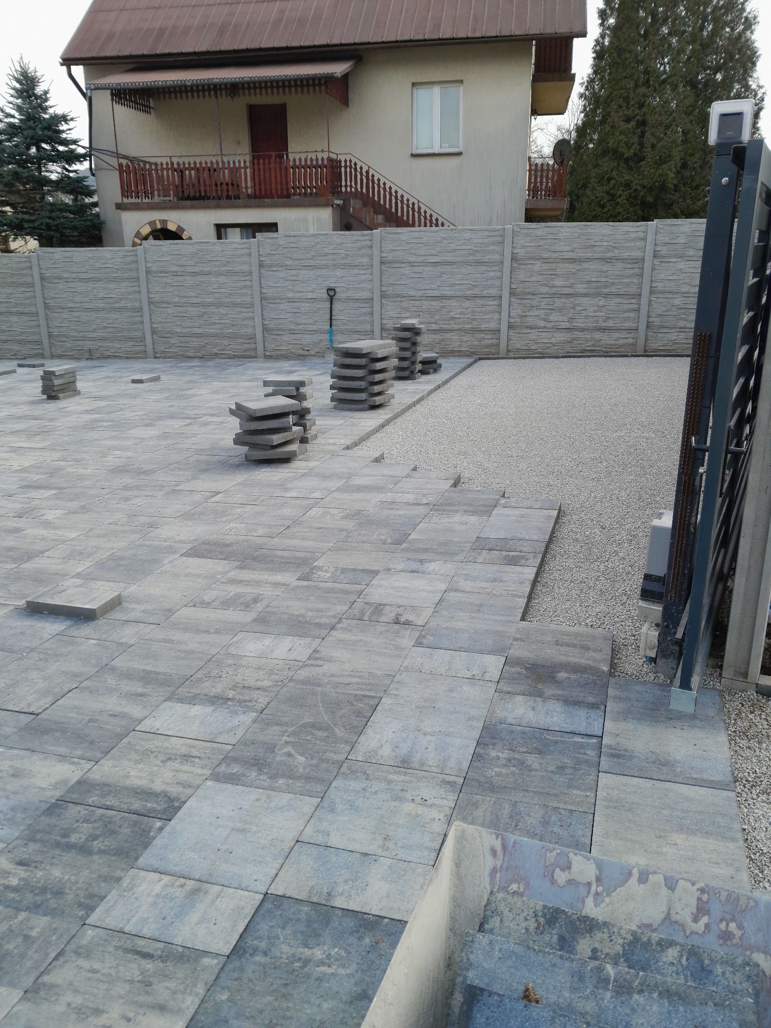 Układanie szarej kostki brukowej na posesji z widocznym ogrodzeniem panelowym i stertami kostki. Częściowo wyłożona powierzchnia, przygotowana pod dalsze prace.