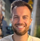 Uśmiechnięty mężczyzna z krótkimi włosami, w białej koszulce, na rozmytym tle z jasnymi punktami świetlnymi. Portret z bliska, naturalny uśmiech.