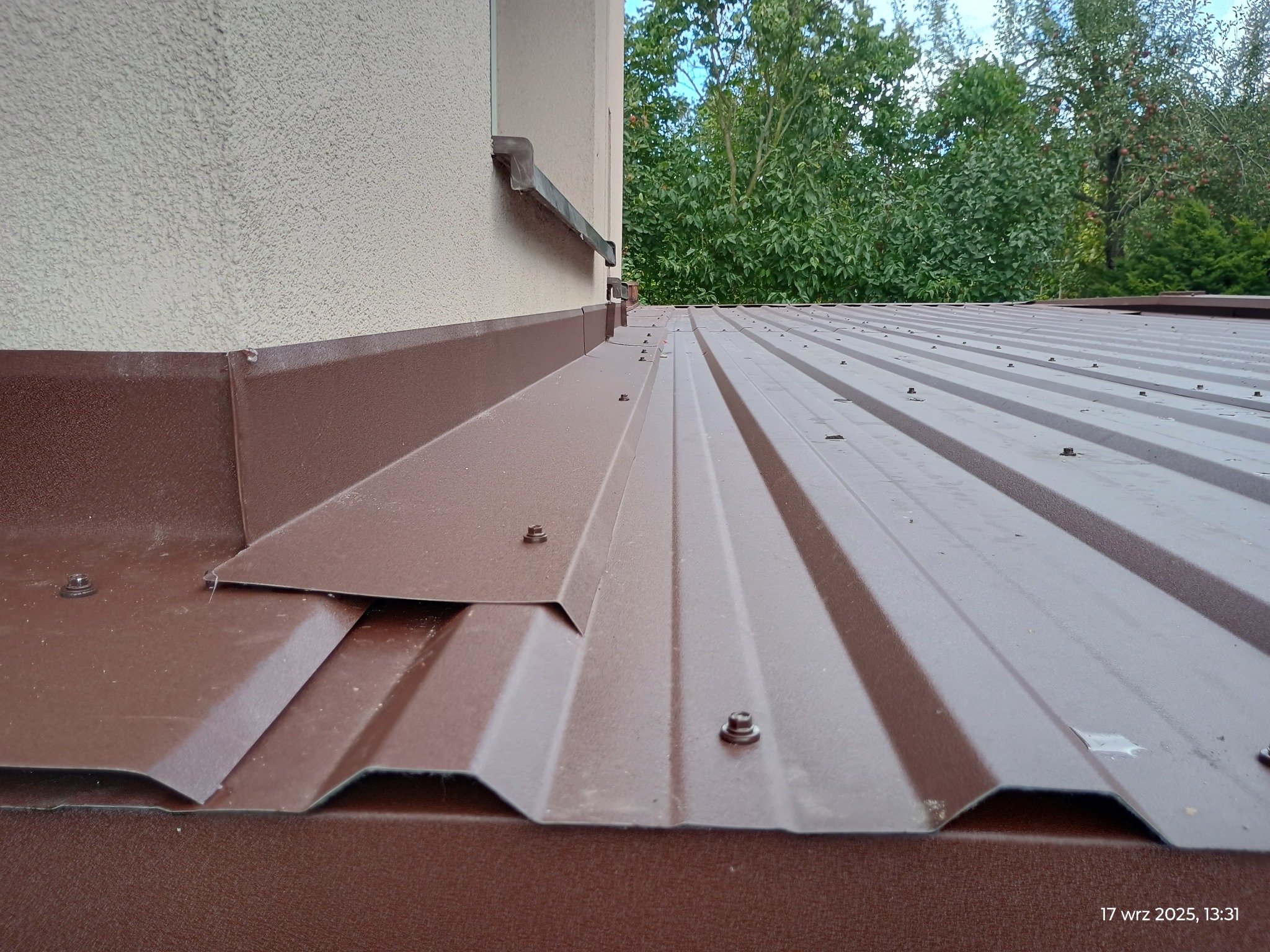 Brązowy dach z blachy trapezowej, mocowany śrubami, z widocznymi obróbkami blacharskimi przy ścianie budynku. Tło stanowią zielone drzewa.