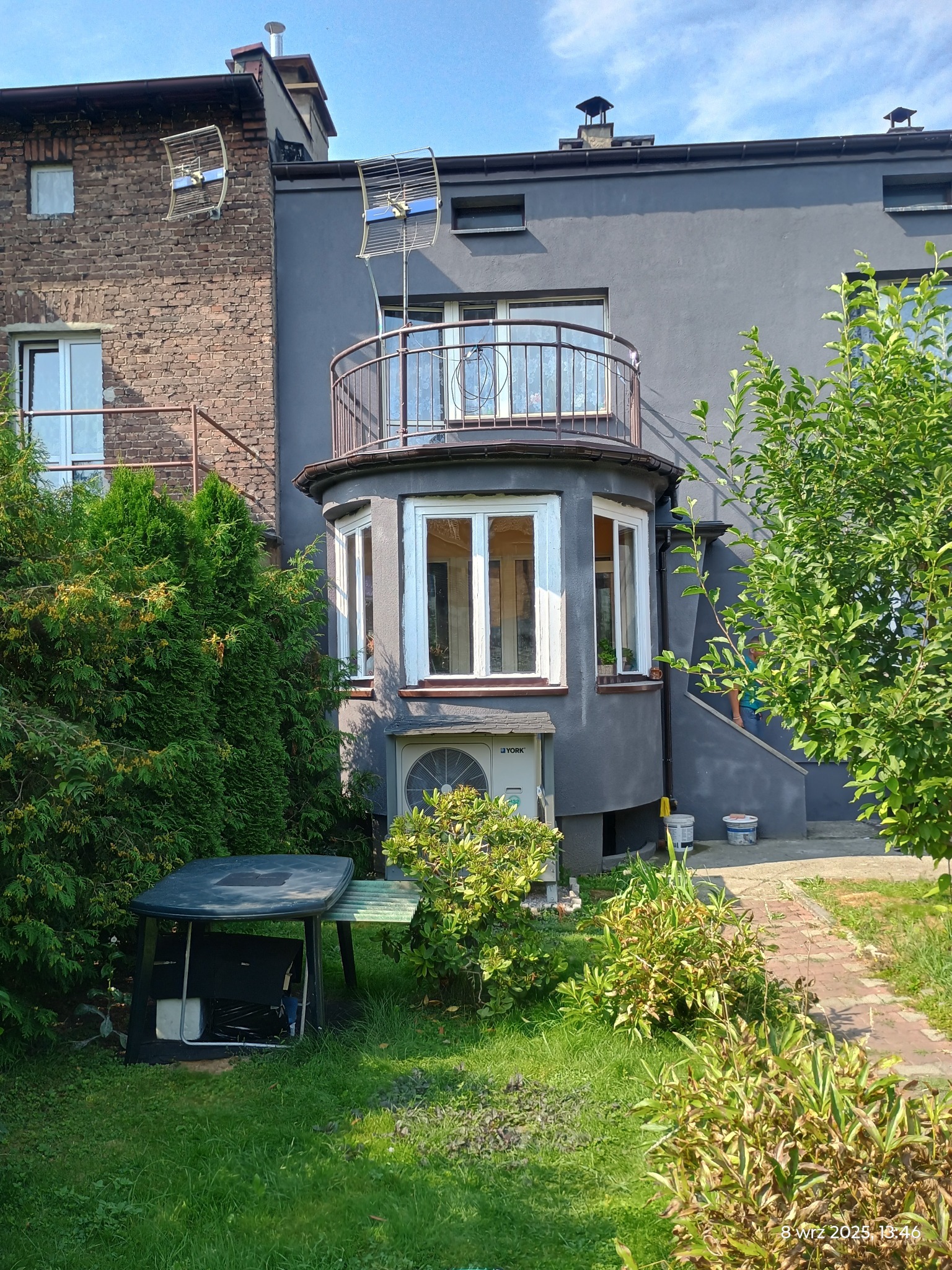 Szary budynek z balkonem i antenami na dachu, widok z ogrodu. Widoczne okna, klimatyzator i schody. Część budynku z cegły. Zieleń w ogrodzie.