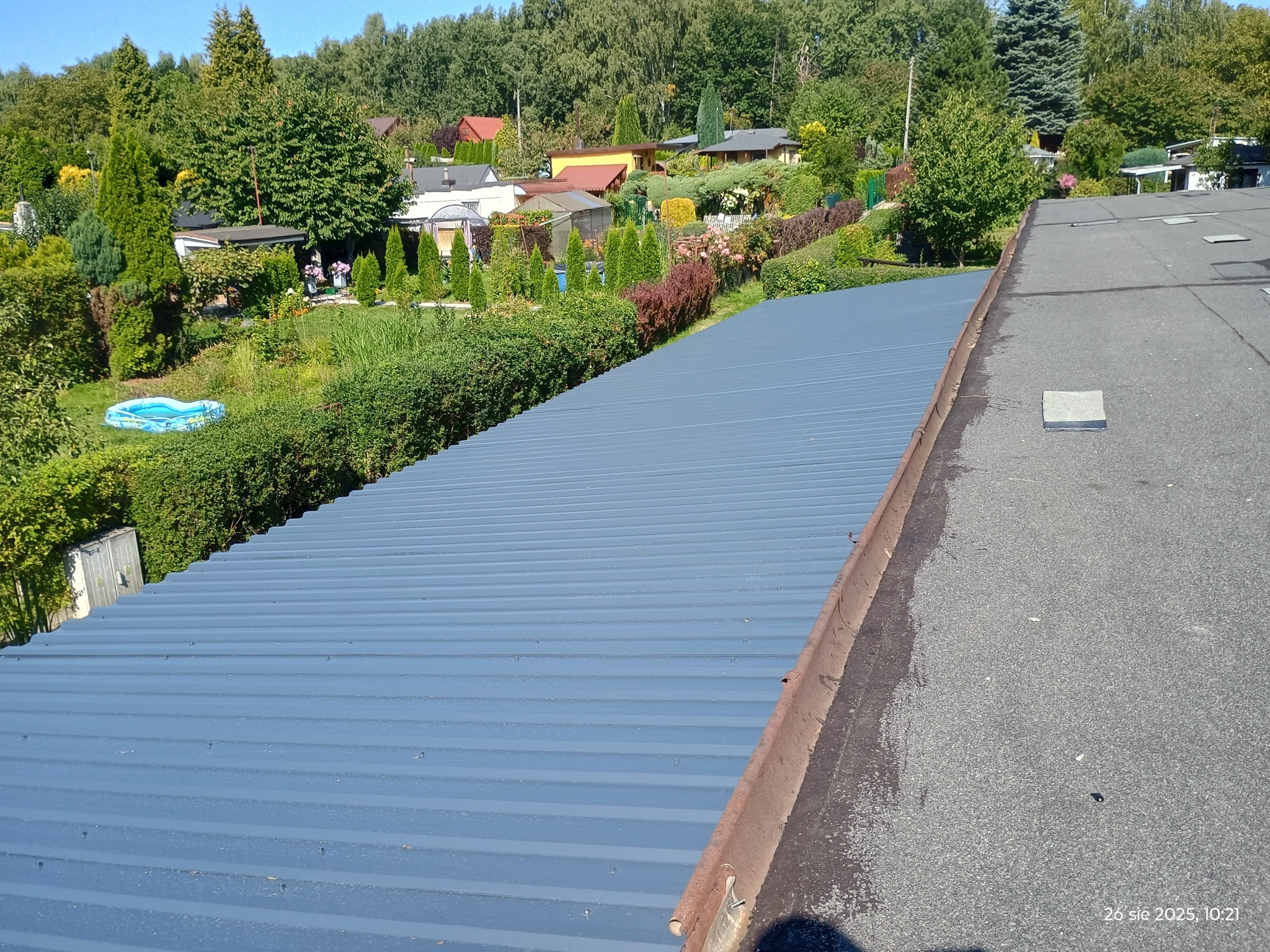 Szary, blaszany dach z widocznymi liniami łączeń, na tle zieleni ogrodów i zabudowań. Ujęcie z góry, ukazujące strukturę dachu i otoczenie.