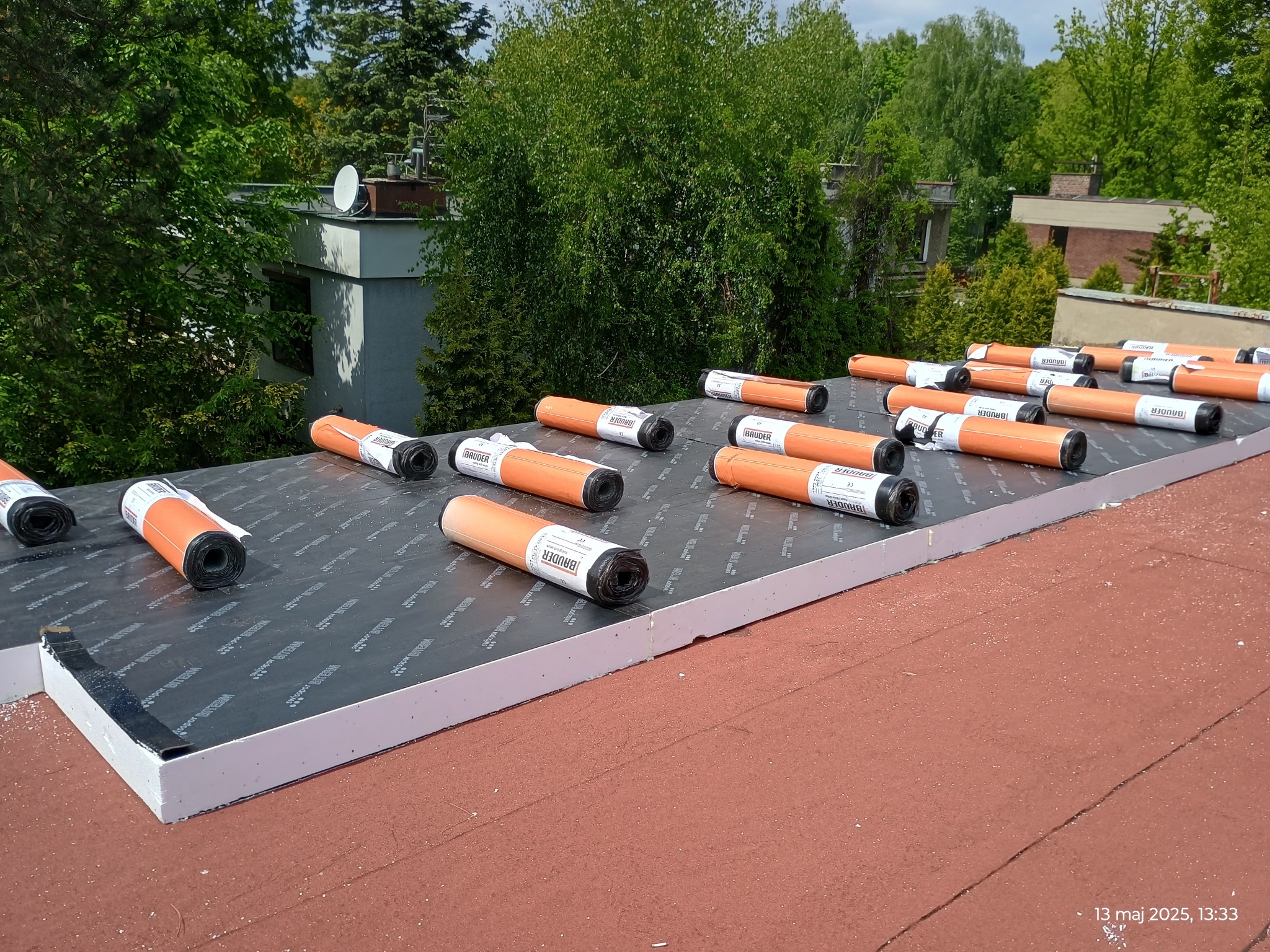Płaski dach z izolacją termiczną i rolkami papy Bauder przed montażem, widok z góry. Białe płyty izolacyjne i pomarańczowe rolki na czarnej membranie dachowej.