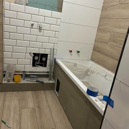 MAR-BUD MARCIN ZAWIŚLIŃSKI - Białe, prostokątne płytki na ścianie w łazience w trakcie montażu, widoczne narzędzia glazurnicze, fragment wanny zabezpieczonej folią i drewnopodobna podłoga.