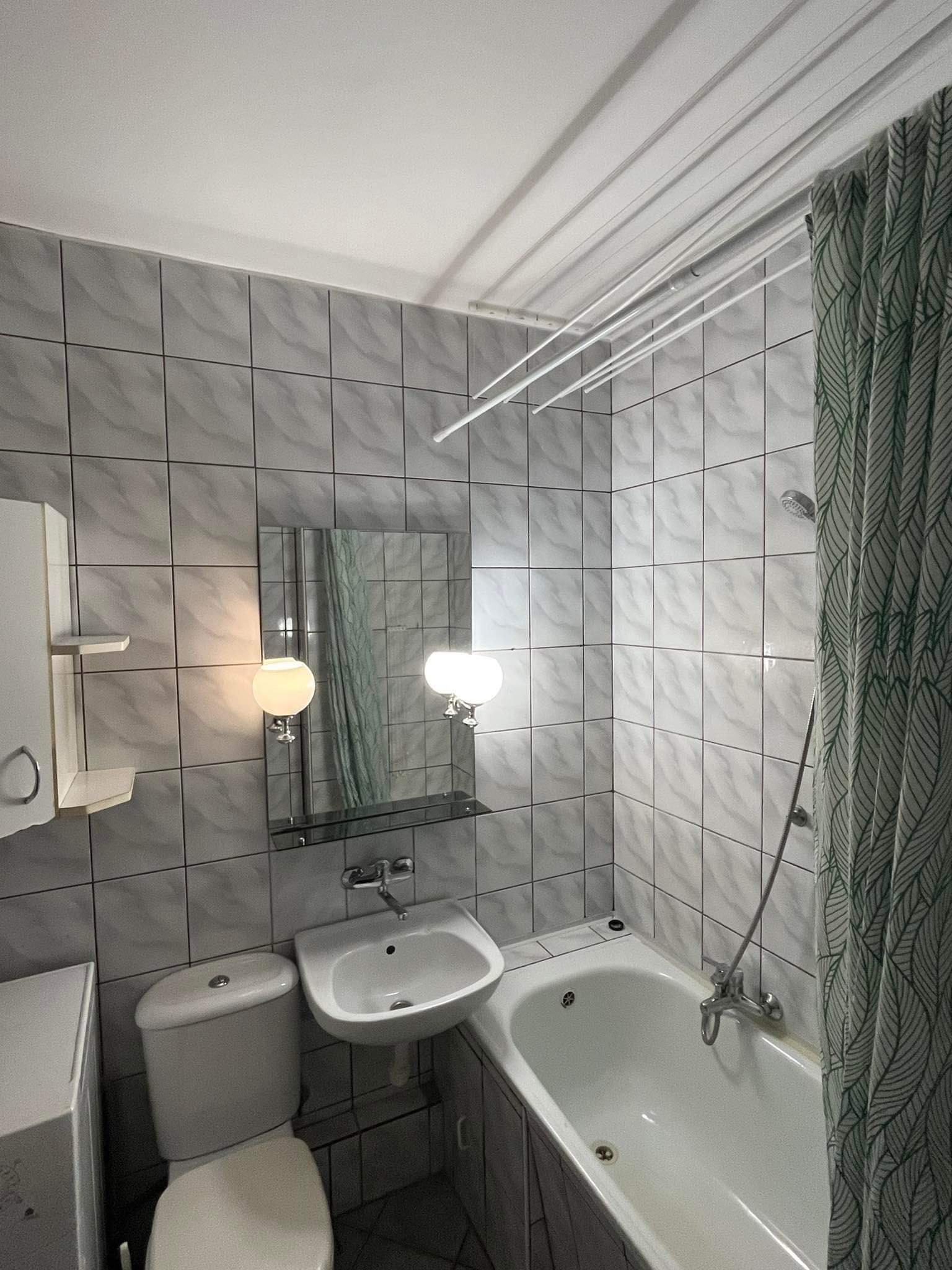 Wykończona łazienka z biało-szarymi płytkami, białą wanną, umywalką i toaletą. Nad umywalką lustro z oświetleniem. Suszarka sufitowa i zasłona w liście.