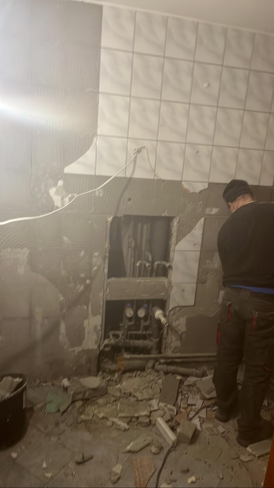 Demontaż płytek w łazience podczas remontu. Widoczne odsłonięte rury i instalacje. Na podłodze gruz i resztki materiałów. Osoba w roboczym ubraniu w tle.