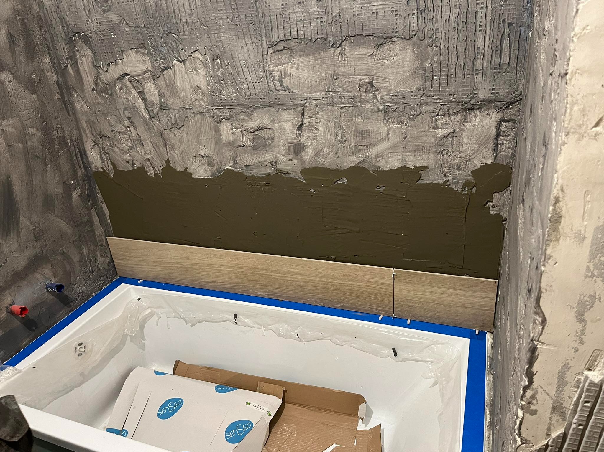 Układanie płytek imitujących drewno nad wanną w łazience. Widoczna warstwa kleju i krzyżyki dystansowe. Ściana w surowym tynku.