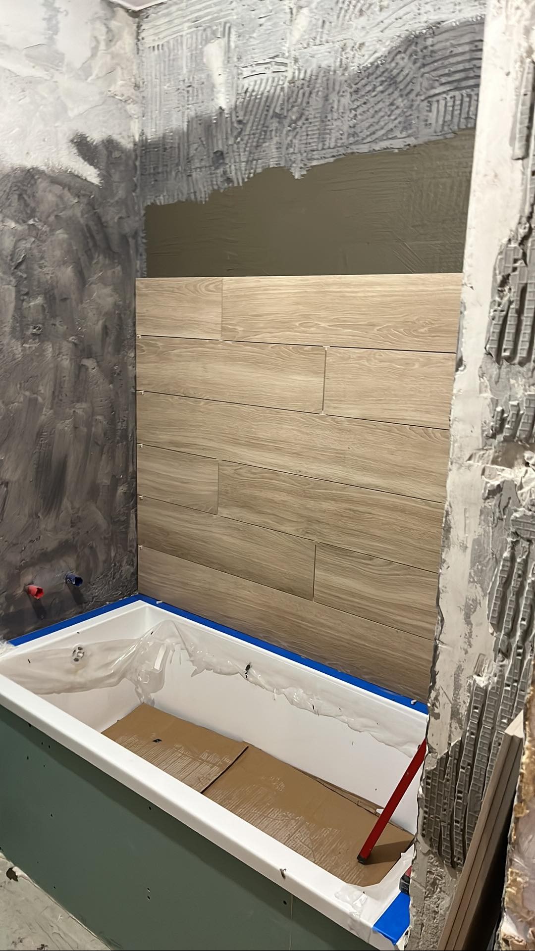 Montaż płytek imitujących drewno na ścianie nad wanną w łazience w trakcie remontu. Widoczne surowe ściany i przygotowane podłoże.