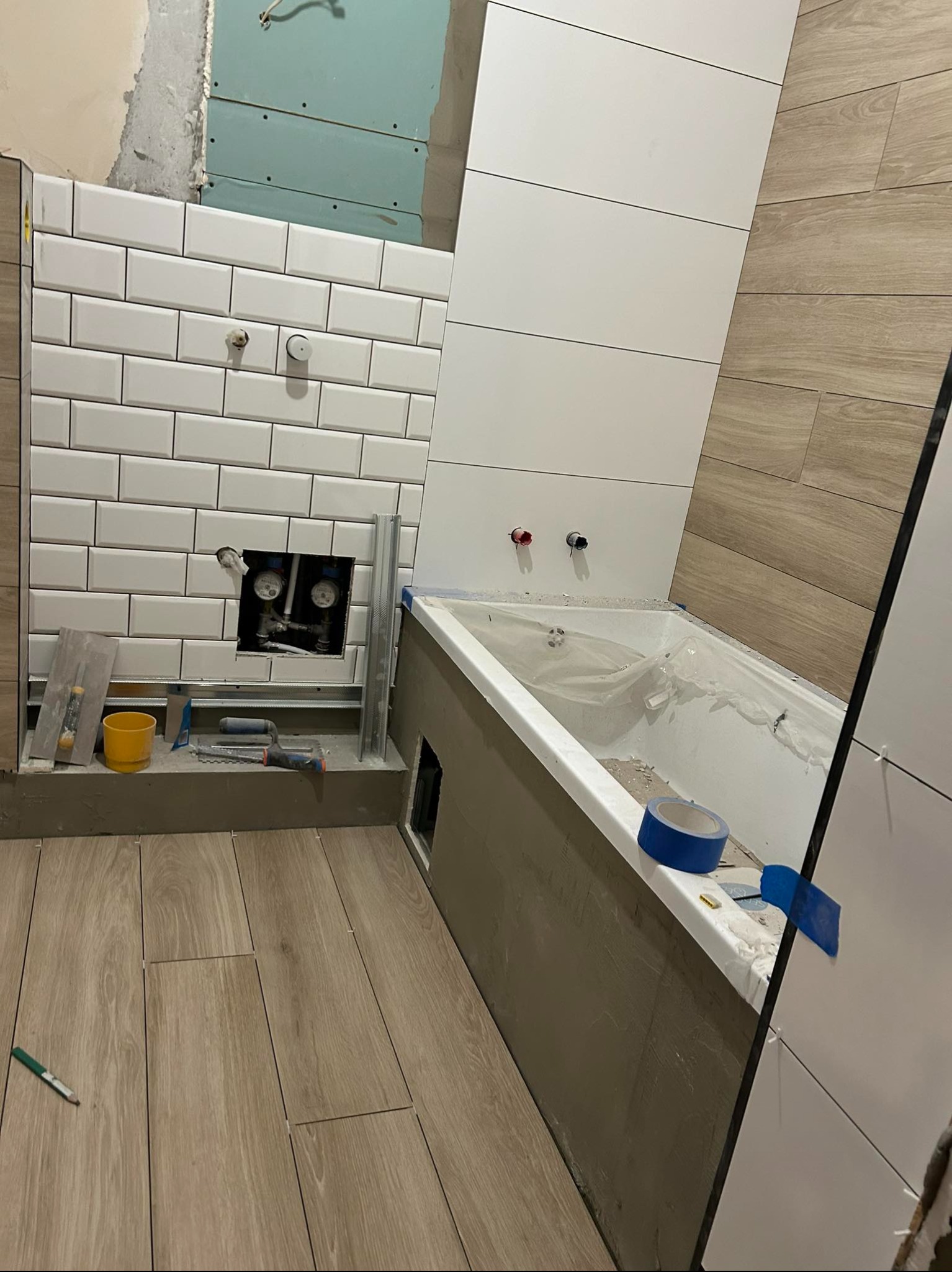 Białe, prostokątne płytki na ścianie w łazience w trakcie montażu, widoczne narzędzia glazurnicze, fragment wanny zabezpieczonej folią i drewnopodobna podłoga.