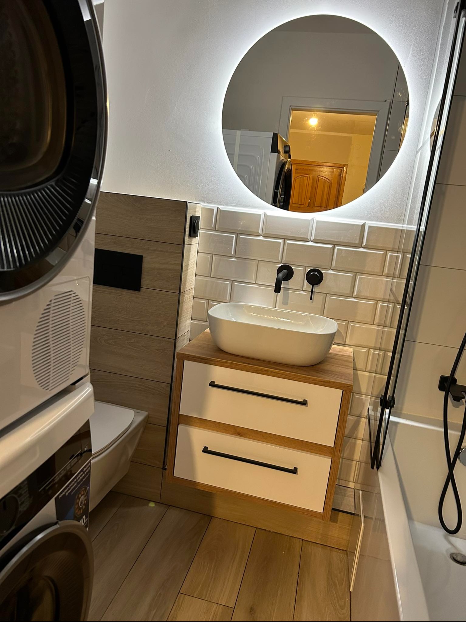 Nowoczesna łazienka z owalną umywalką nablatową, szafką z szufladami, okrągłym lustrem z podświetleniem LED i białą armaturą. Widoczna pralka.