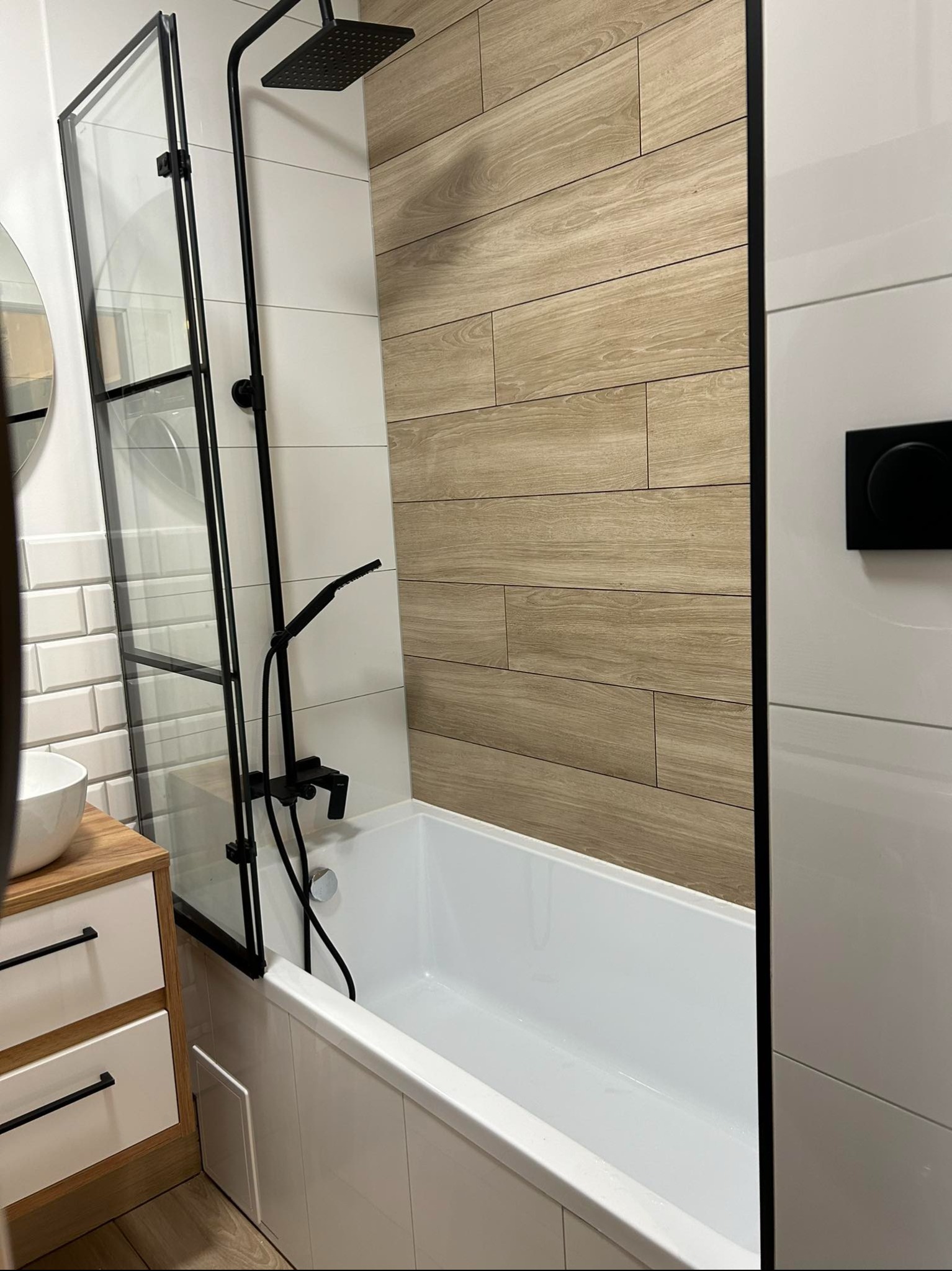 Nowoczesna łazienka z wanną, czarną armaturą i drewnopodobnymi płytkami. Kabina prysznicowa z czarnymi profilami. Umywalka z szafką w stylu skandynawskim.