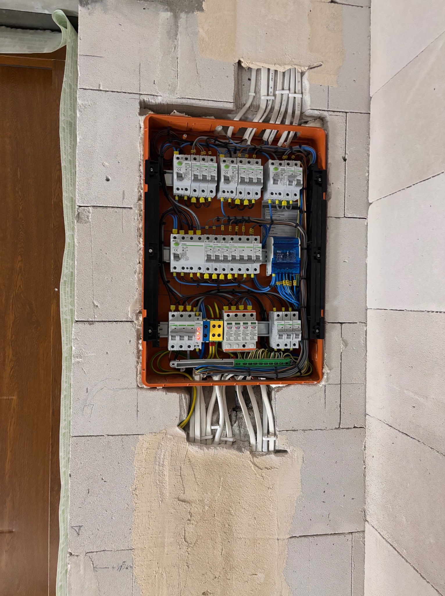 Pomarańczowa skrzynka z bezpiecznikami i przewodami w surowej ścianie z betonu komórkowego. Instalacja elektryczna w trakcie montażu, widoczne kable i zabezpieczenia.