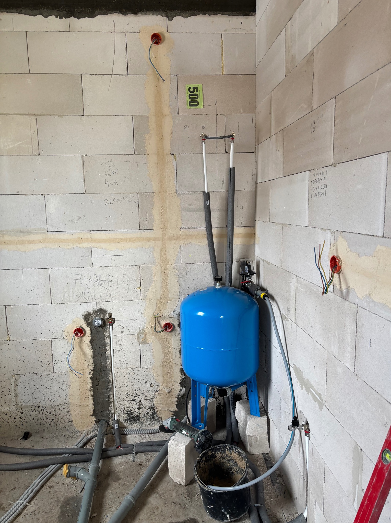 Niebieski hydrofor w surowym wnętrzu z instalacjami: elektryczną i hydrauliczną. Widoczne przewody, puszki elektryczne i fragment ściany z bloczków.