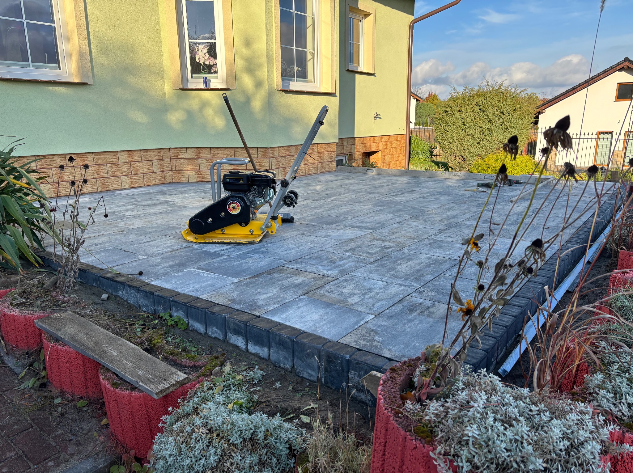 Ułożona kostka brukowa na posesji w Roszkowie, z żółtą zagęszczarką w tle. Widoczne donice i fragment domu. Kąt ujęcia z dołu podkreśla równość powierzchni.