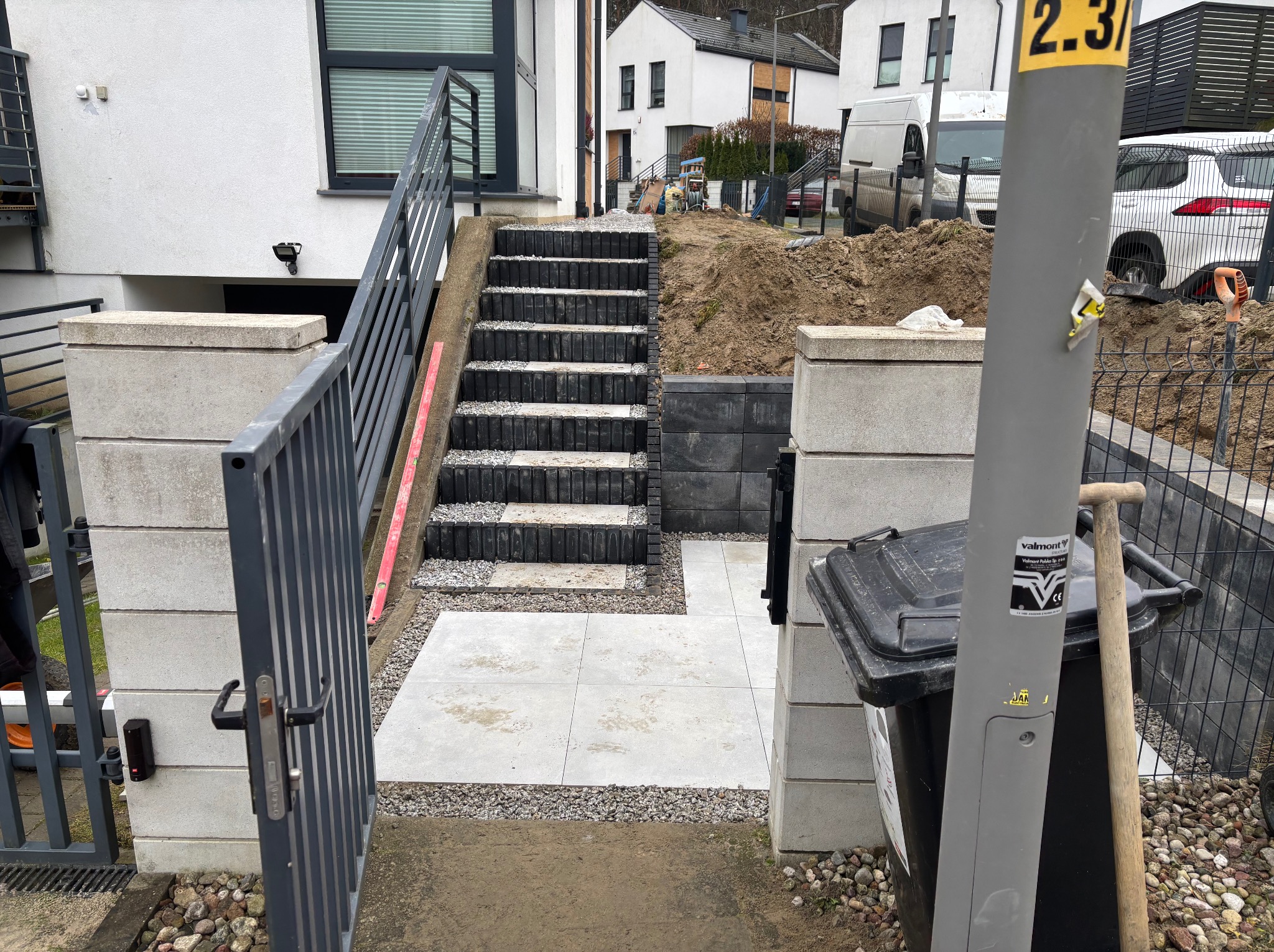 Nowe schody i chodnik z szarej kostki prowadzące do domu, widok zza otwartej furtki. W tle dom z białymi ścianami i czarnymi oknami, ziemia i materiały budowlane.