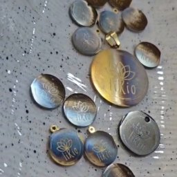 NOIR - Metalowe, okrągłe zawieszki z wygrawerowanym logo i motywem kwiatowym, w różnych odcieniach złota, srebra i niebieskiego, leżące na nakrapianej powierzchni.