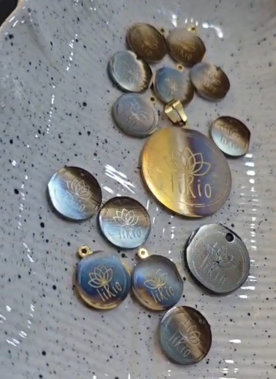 Metalowe, okrągłe zawieszki z wygrawerowanym logo i motywem kwiatowym, w różnych odcieniach złota, srebra i niebieskiego, leżące na nakrapianej powierzchni.