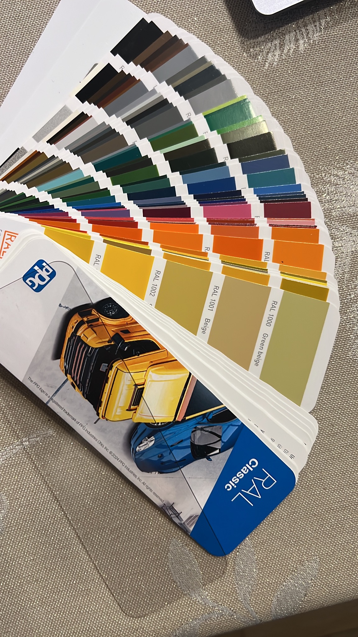 Paleta kolorów RAL Classic z oznaczeniami odcieni, prezentująca szeroki wachlarz barw od szarości i zieleni po pomarańcze i róże, z logo PPG i ilustracjami pojazdów.