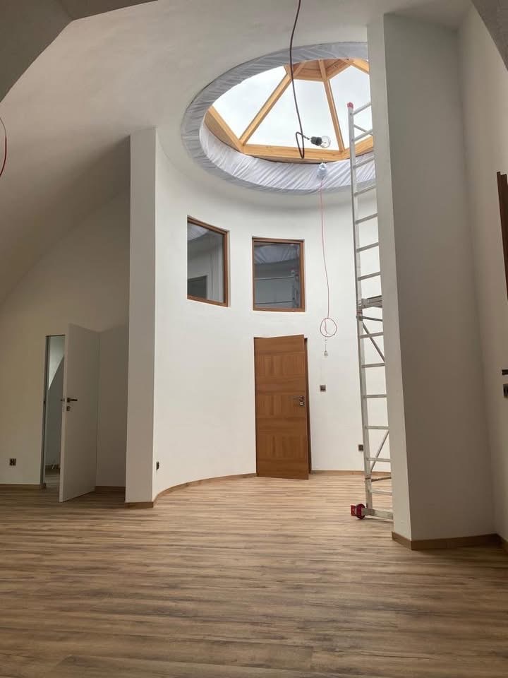 Jasne wnętrze z drewnianą podłogą i oknem dachowym. Ściany białe, częściowo zaokrąglone. Drabina oparta o ścianę. Widoczne drzwi i okna.