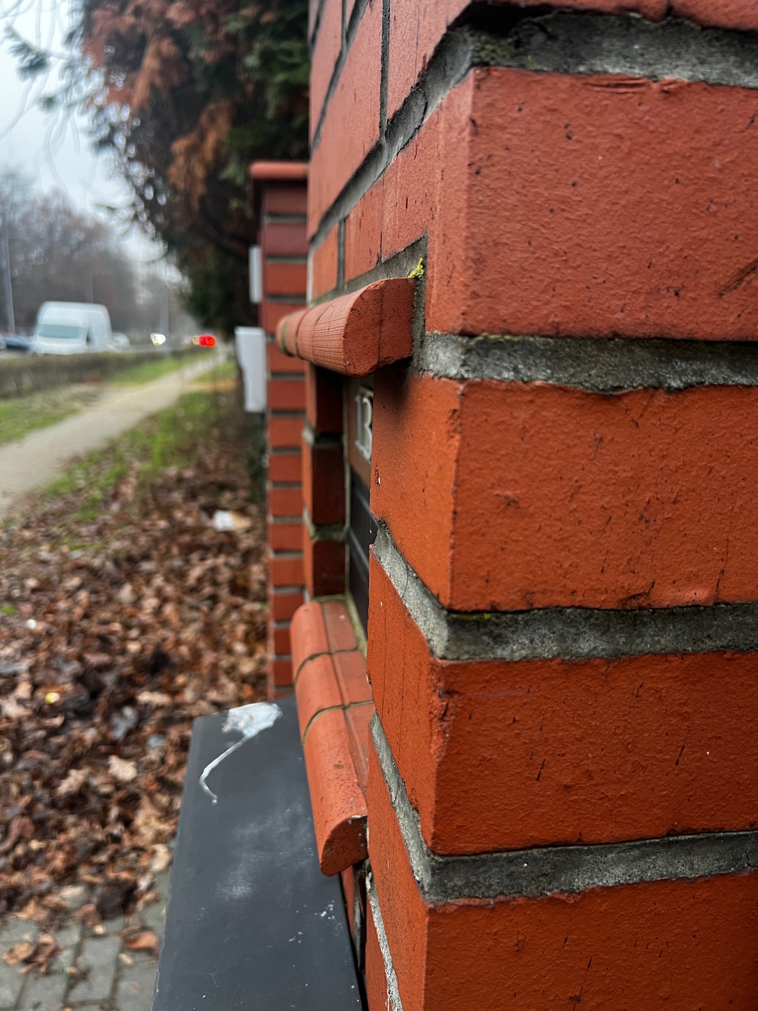 Ceglany fragment ogrodzenia z dekoracyjnymi elementami, widok z boku, z liśćmi na ziemi i ulicą w tle. Detal architektury ogrodowej w pochmurny dzień.