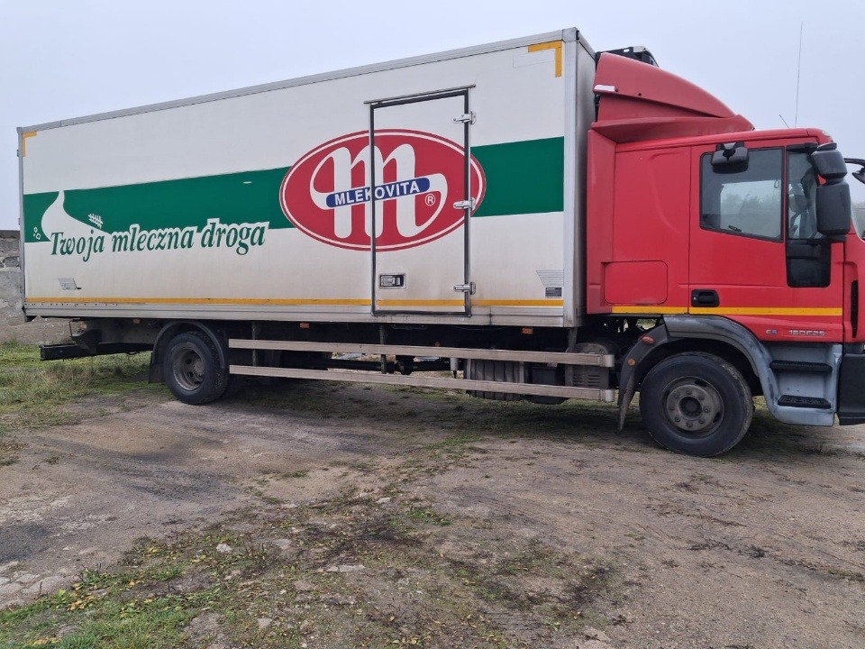 Czerwono-biała ciężarówka chłodnia z logo Mlekovita na naczepie, zaparkowana na szutrowym podłożu. Widok z boku, pochmurne niebo w tle.