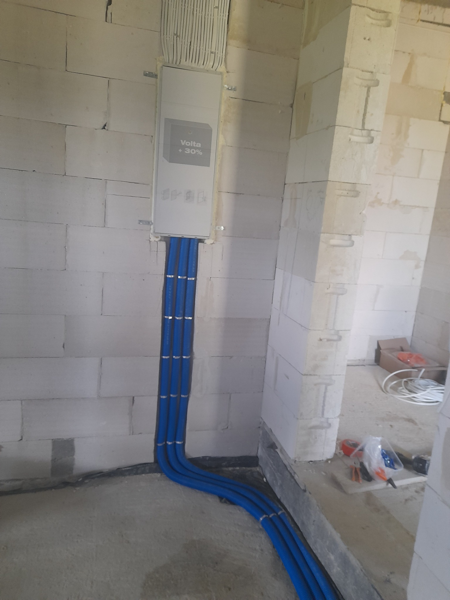 Rozdzielnia elektryczna Volta +30% z trzema niebieskimi rurami instalacyjnymi w surowym wnętrzu budynku z białych bloczków. Widok na podłogę i fragment ściany.