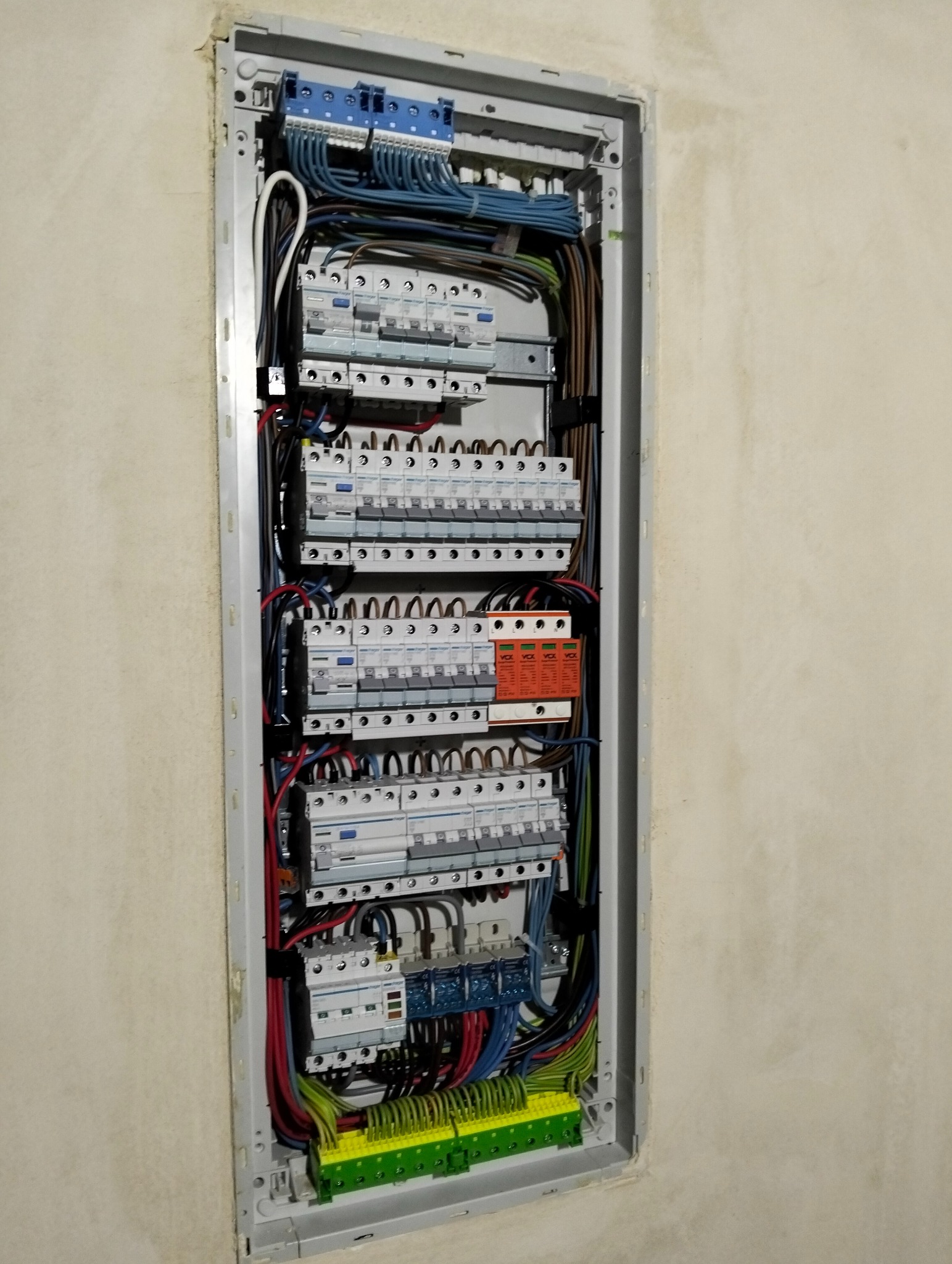 Rozdzielnica elektryczna wnękowa z zainstalowanymi bezpiecznikami, przewodami i listwami. Widoczne kable w różnych kolorach. Ściana w tle w jasnym odcieniu.