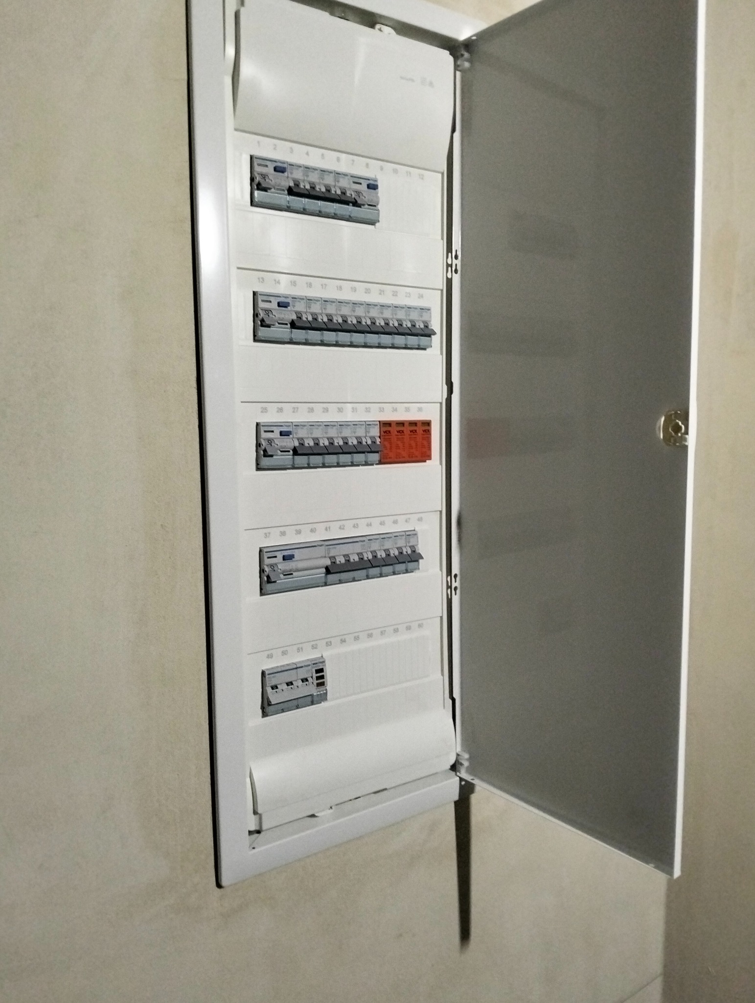 Otwarta, biała skrzynka bezpiecznikowa z numerowanymi wyłącznikami, w tym pomarańczowe. Widok z bliska na ścianie, prezentujący instalację elektryczną.