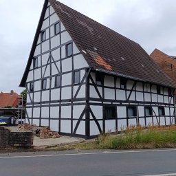 Gnat Bud - Odnowiony dom szachulcowy z ciemnymi belkami na białej fasadzie, widok z ulicy, z fragmentem rozbiórki muru w tle i elementami rusztowania.