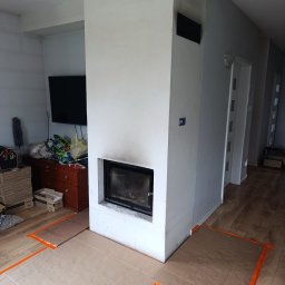 Gnat Bud - Wnętrze w trakcie prac wykończeniowych: obudowa kominka z kratkami wentylacyjnymi, zabezpieczona podłoga kartonem i taśmą. W tle widoczny telewizor i komoda.