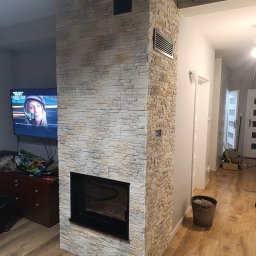 Gnat Bud - Obudowa kominka z dekoracyjnej, jasnej cegły w salonie z drewnianą podłogą. Widoczny fragment telewizora i drzwi w tle. Wykończenie wnętrza.