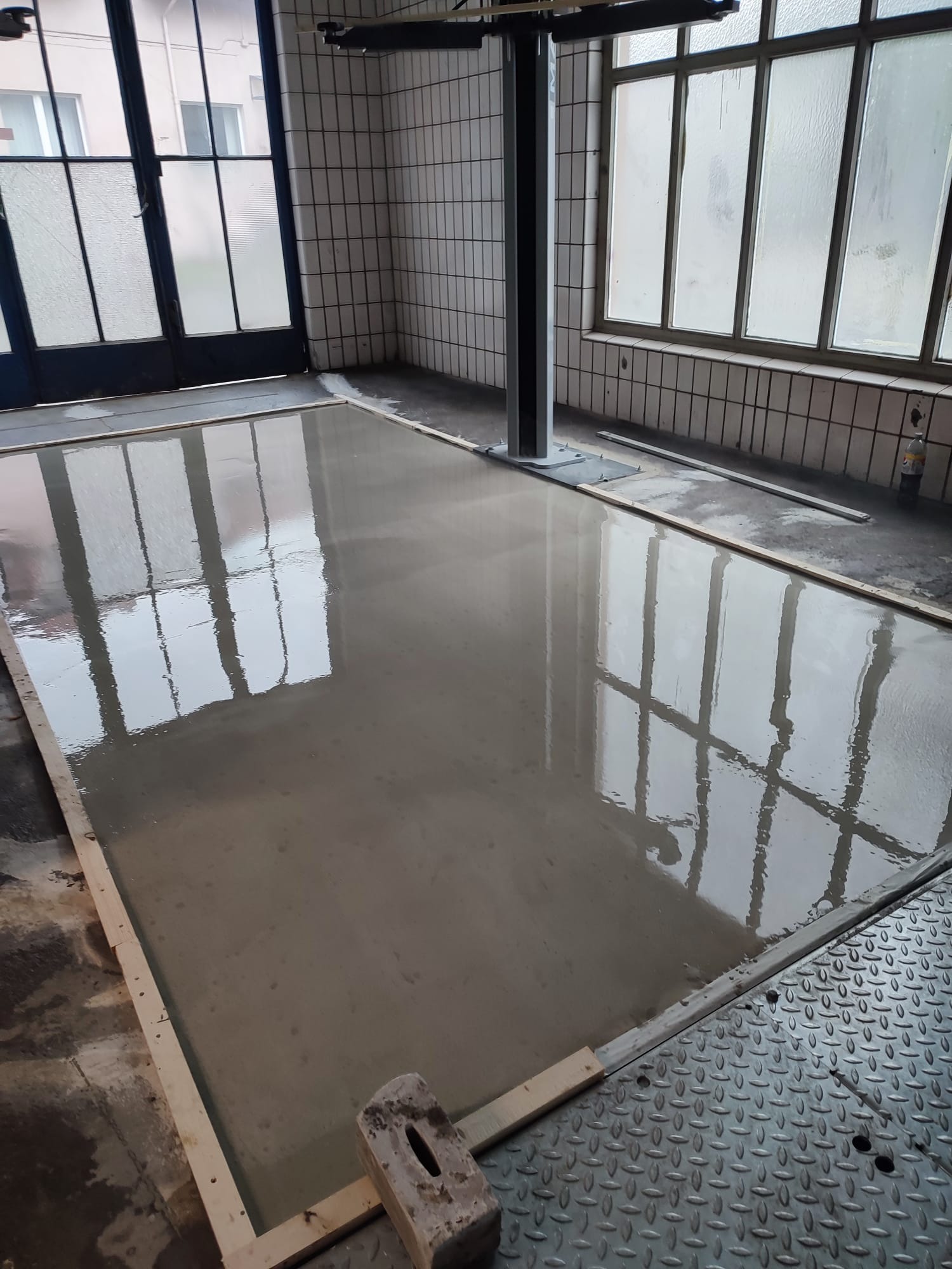 Wylana, świeża posadzka w pomieszczeniu z oknami i białą, kwadratową glazurą na ścianach. Widoczne drewniane obramowanie i młotek. Refleksy okien na mokrej powierzchni.