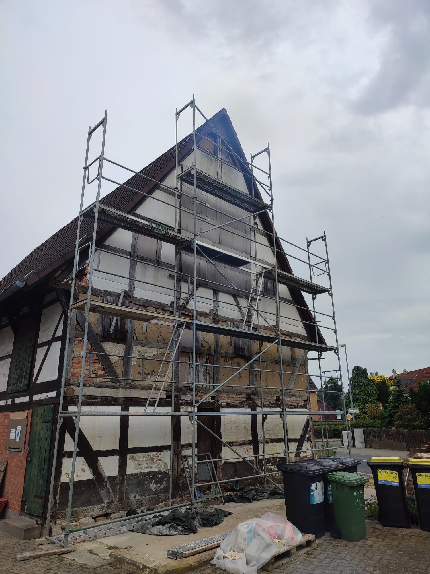 Dom szkieletowy w trakcie renowacji, otoczony rusztowaniem. Widoczne uszkodzenia elewacji i przygotowania do prac remontowych. Ujęcie z dołu, pochmurne niebo w tle.