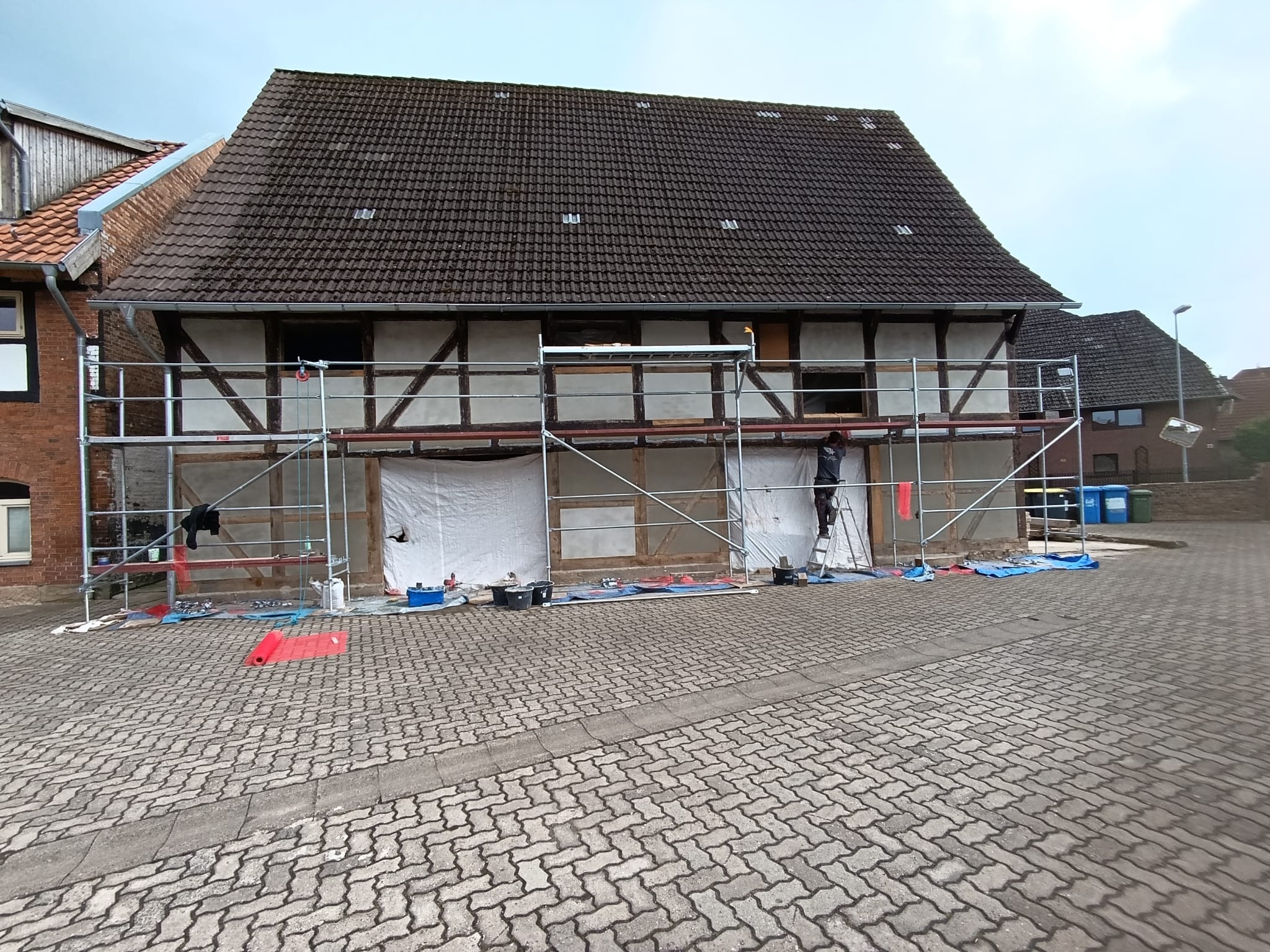 Renowacja elewacji domu szkieletowego z rusztowaniem. Prace w toku, zabezpieczone okna, widoczne narzędzia i materiały budowlane. Brukowana droga na pierwszym planie.