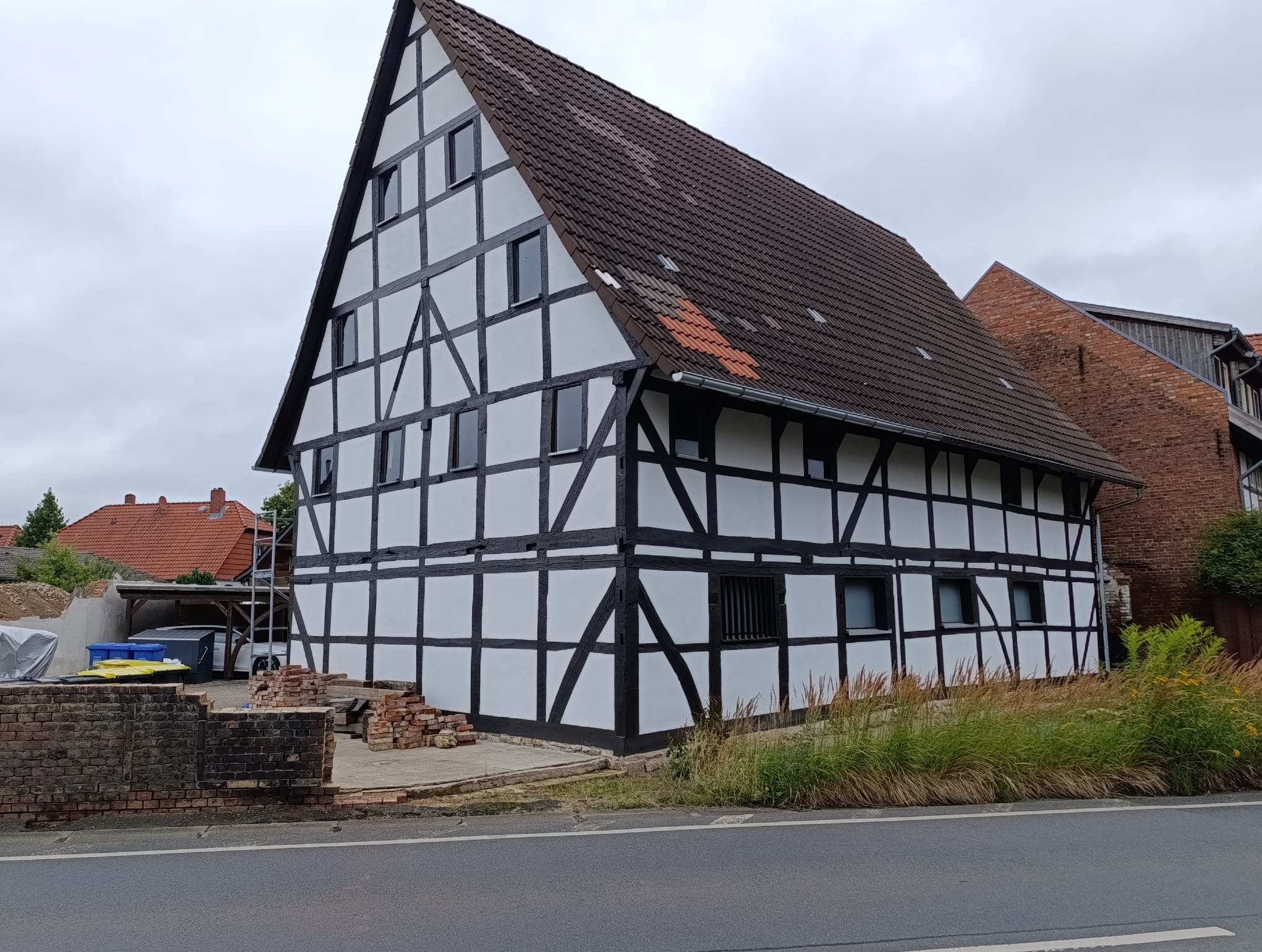 Odnowiony dom szachulcowy z ciemnymi belkami na białej fasadzie, widok z ulicy, z fragmentem rozbiórki muru w tle i elementami rusztowania.