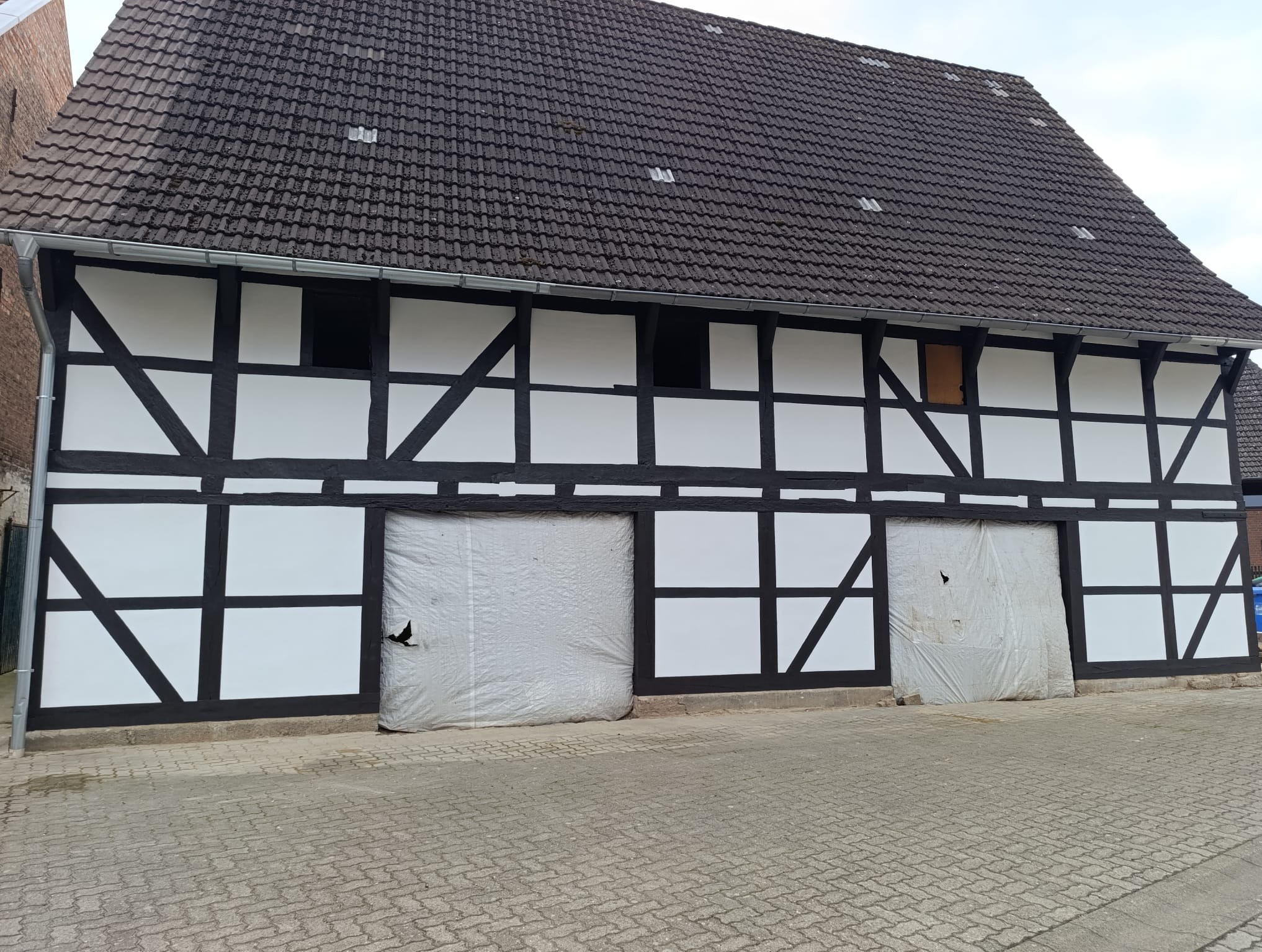 Szachulcowy budynek z ciemną dachówką, zabezpieczone wejścia folią. Widoczne czarno-białe ramy konstrukcyjne i fragment brukowanej drogi. Ujęcie z dołu, pochmurne niebo.