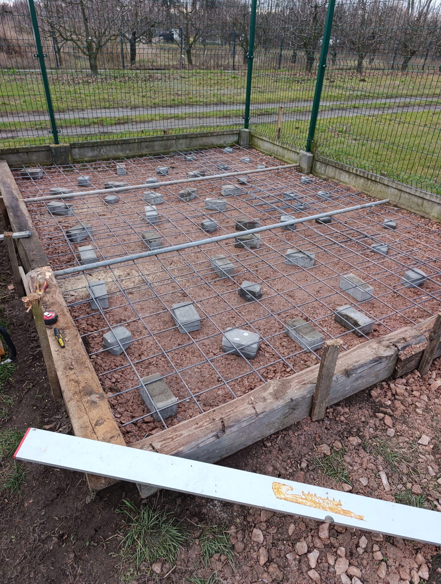 Przygotowanie podłoża pod wylewkę: szalunek z desek, siatka zbrojeniowa podniesiona kostkami, poziomica w kadrze. Widok z góry na plac budowy.