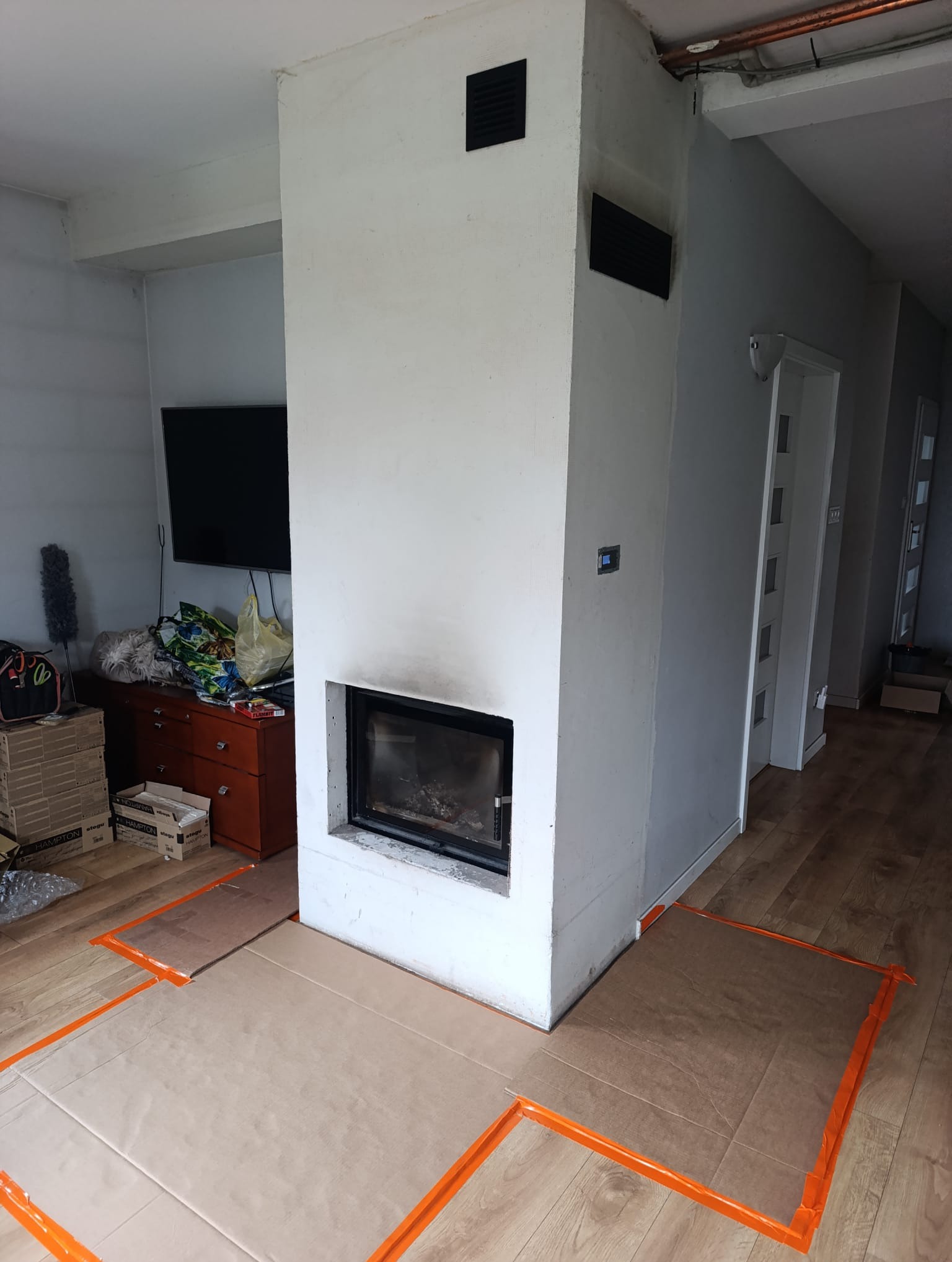 Wnętrze w trakcie prac wykończeniowych: obudowa kominka z kratkami wentylacyjnymi, zabezpieczona podłoga kartonem i taśmą. W tle widoczny telewizor i komoda.