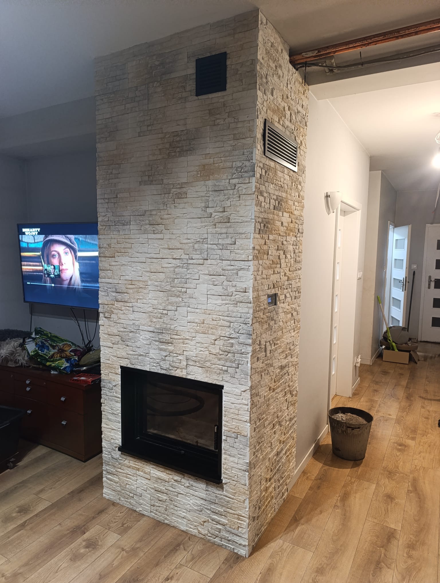 Obudowa kominka z dekoracyjnej, jasnej cegły w salonie z drewnianą podłogą. Widoczny fragment telewizora i drzwi w tle. Wykończenie wnętrza.