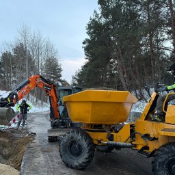 Euro-Standard - Wynajem Maszyn - Roboty Ziemne i Budowlane - Kruszywa - Transport - Pomarańczowa koparka i żółta wywrotka na tle wykopu i drzew. Operator w kamizelce. Widoczny znak stacji benzynowej w oddali. Praca w toku, pochmurne niebo.