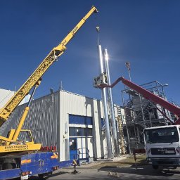 Euro-Standard - Wynajem Maszyn - Roboty Ziemne i Budowlane - Kruszywa - Transport - Montaż komina przemysłowego z użyciem dźwigu i podnośnika koszowego na tle hali produkcyjnej w słoneczny dzień. Widoczny pracownik w koszu podnośnika.