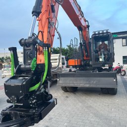 Euro-Standard - Wynajem Maszyn - Roboty Ziemne i Budowlane - Kruszywa - Transport - Pomarańczowa koparka z czarnym osprzętem na placu, operator w kabinie. Widoczne detale: hydraulika, logo 'STFELWRIST X20'. W tle budynek firmy i inne pojazdy.