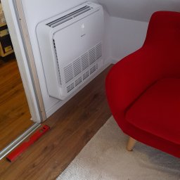 Martin Serwis - Biała klimatyzacja Hisense zamontowana na skośnej ścianie poddasza, obok czerwonego fotela i poziomicy na podłodze. Widoczny fragment puszystego dywanu.