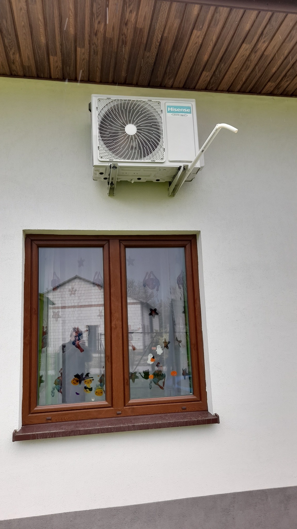 Biały klimatyzator Hisense zamontowany na elewacji domu nad oknem z dekoracjami Halloween. Widoczne rury odprowadzające skropliny i drewniana podbitka dachu.