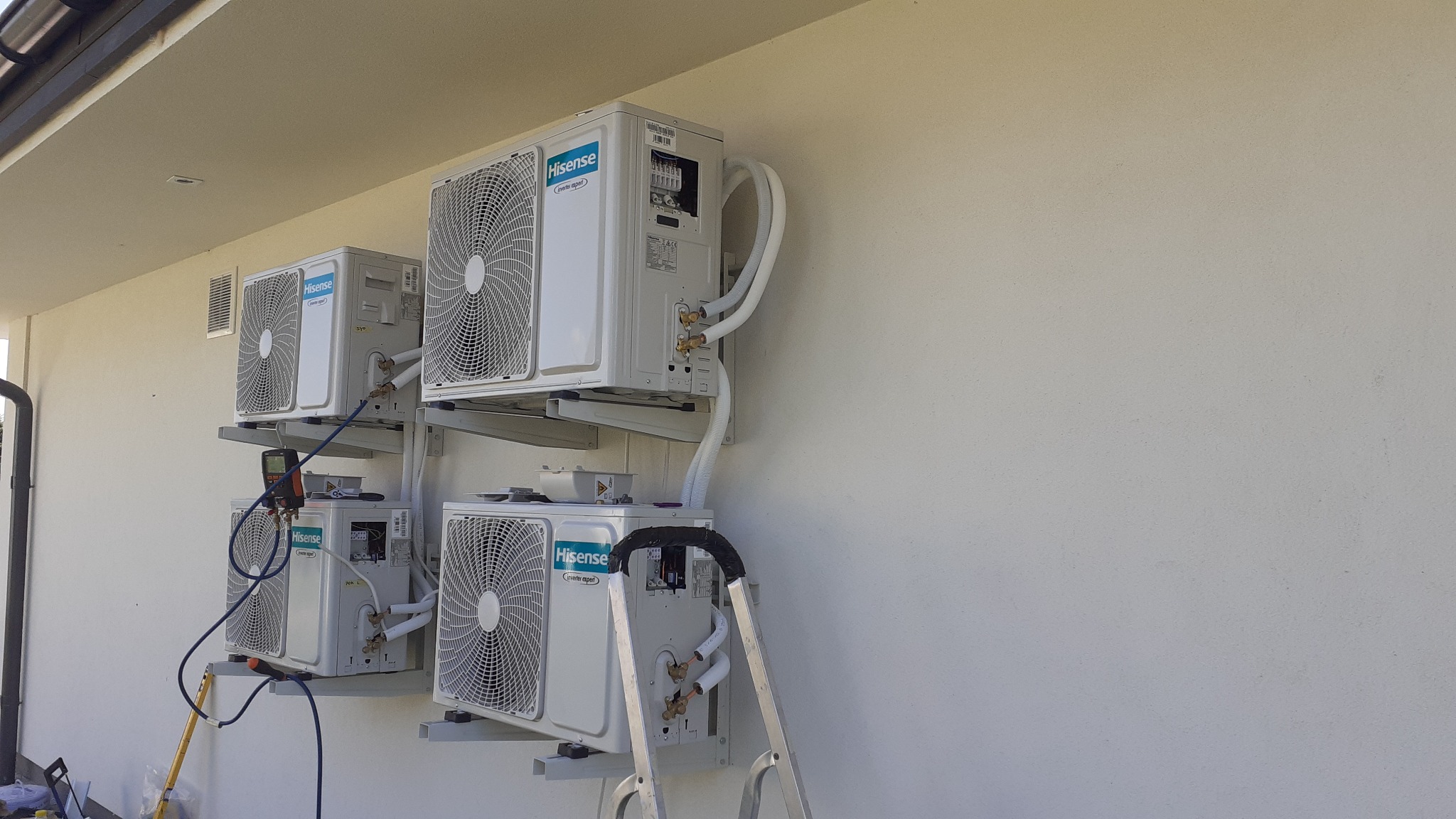 Cztery klimatyzatory Hisense zamontowane na zewnątrz budynku, połączone rurami. Widoczna drabina i narzędzia, sugerujące trwający montaż lub serwis.