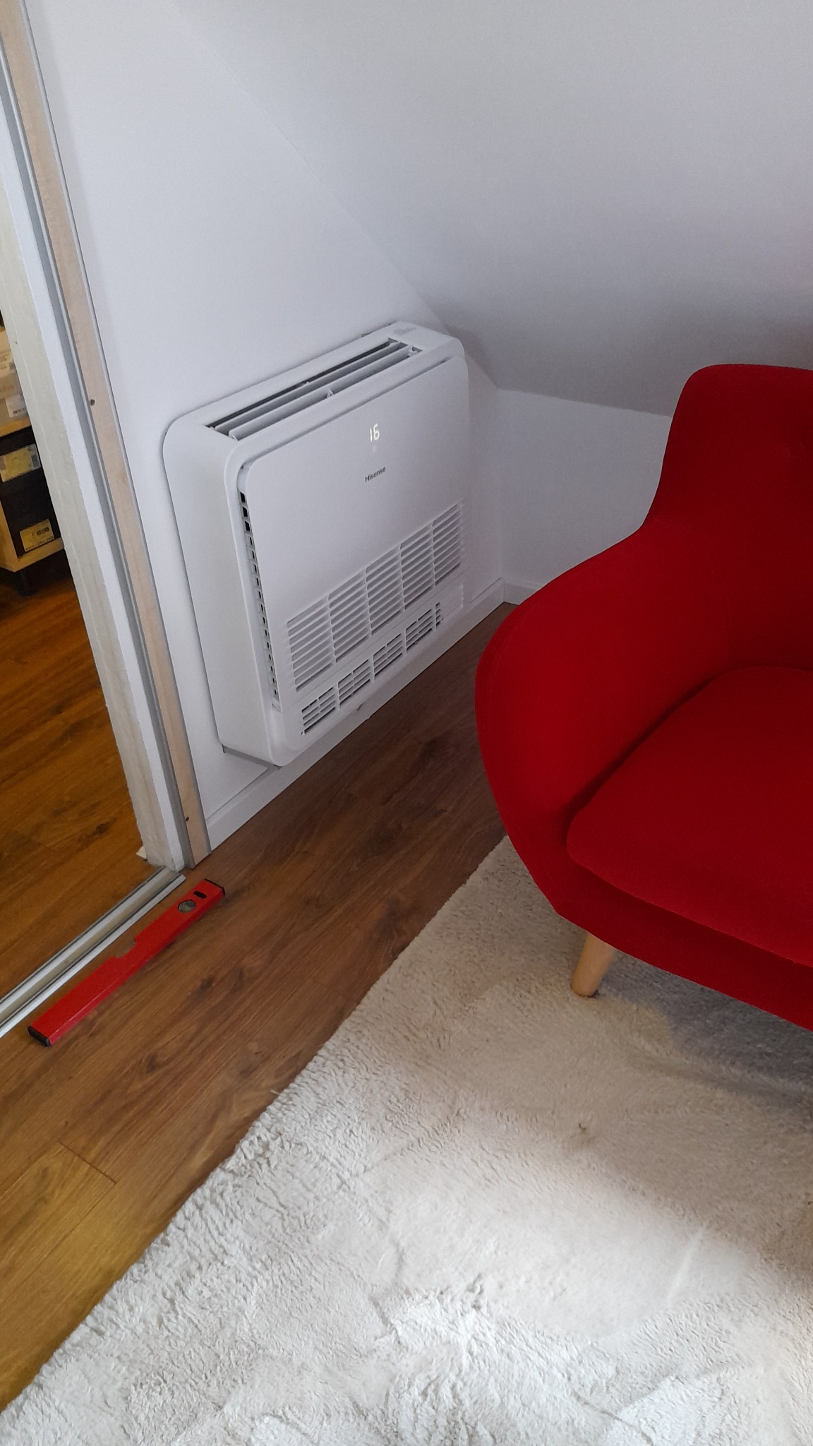 Biała klimatyzacja Hisense zamontowana na skośnej ścianie poddasza, obok czerwonego fotela i poziomicy na podłodze. Widoczny fragment puszystego dywanu.