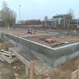 USŁUGI OGÓLNOBUDOWLANE - KAMIL KOBIELA - Zalany fundament pod budynek w Skrzeszewie Żukowskim. Widoczne szalunki i deski. Świeży beton, ziemia i drzewa w tle. Ujęcie z bliska, pochmurny dzień.