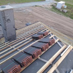 USŁUGI OGÓLNOBUDOWLANE - KAMIL KOBIELA - Układanie dachówki ceramicznej na konstrukcji więźby dachowej. Widoczny komin z izolacją i przygotowane pakiety dachówek. Tło: pole i droga gruntowa.
